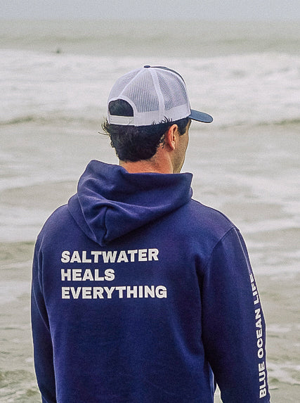 Person wearing a navy blue hoodie with 'Saltwater Heals Everything' text on a beach.